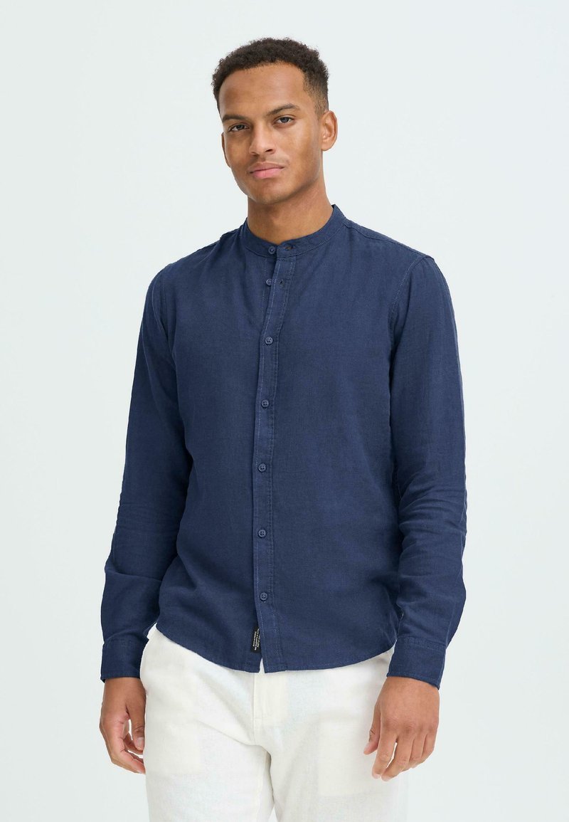 Νέος άνδρας που φοράει μπλε πουκάμισο με στενή γιακά και κουμπιά και άσπρο παντελόνι, στέκεται μπροστά σε απλό φωτεινό φόντο.