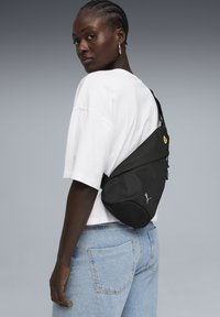 Sac bandoulière noir en matériau textur é, avec un logo jaune et une sangle réglable. Porté sur le corps, en complément d'une chemise blanche.