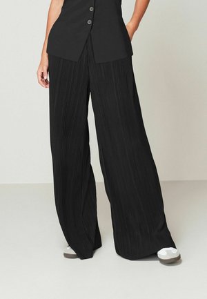 Pantalon large noir plissé avec une finition texturée, associé à un haut noir ajusté avec des boutons. Chaussures décontractées partiellement visibles.