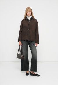 Bruine button-up jas met een gestructureerd ontwerp, gecombineerd met een geruite blouse, grijze wijde jeans en zwarte ballerina's, terwijl ze een kleine donkere handtas vasthoudt.
