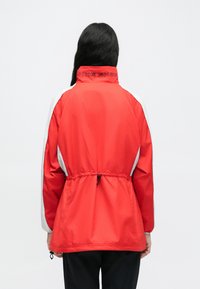 Jachetă roșie din nylon cu accente albe, talie cu șnur, guler înalt și logo "NIKE SHOX" pe spate. Textură netedă și design sportiv.