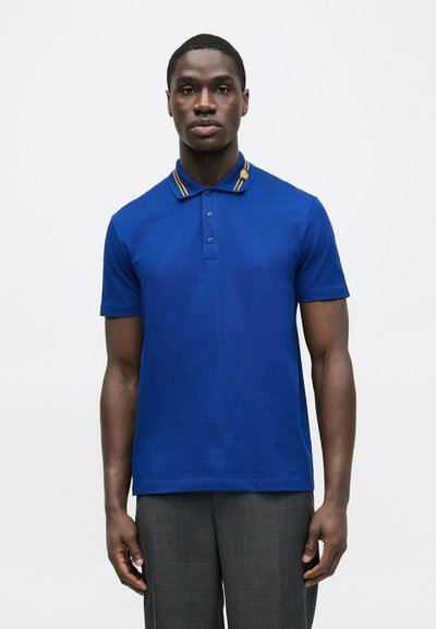 Mėlyna polo marškinėliai trumpomis rankovėmis ir vientisa tekstūra. Yra apykaklė su geltonomis juostelėmis ir mažas emblemėlė kairėje pusėje.