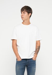 Camiseta de algodón blanca con cuello redondo y mangas cortas, presentada con un corte holgado. Se lleva con jeans de mezclilla azul, mostrando un tatuaje en el brazo.