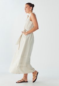 Dlhé, bezrukávové šaty vyrobené z ľahkej béžovej tkaniny, s opaskom v oblasti pása a bočnými vreckami, s vrstveným spodným dielom a jemnou textúrou.