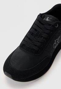 Černá sportovní bota s kombinací hladkého nylonu a texturovaného semiše. Má černé tkaničky a stříbrné logo. Kulatá špička a polstrovaná podrážka.