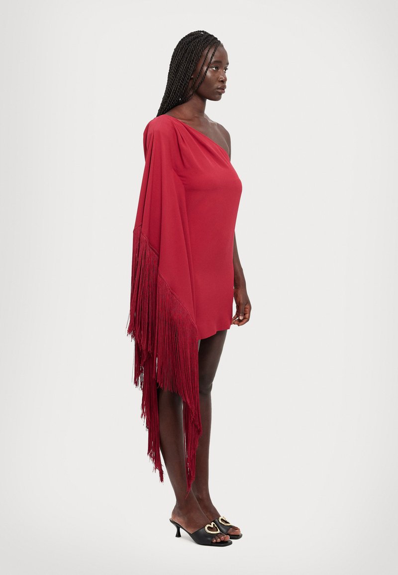Vestido rojo de un solo hombro con una silueta fluida y detalles de flecos largos. Estilizado con mules negros con acentos en forma de corazón.