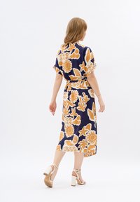 Robe à motifs en bleu marine avec de grands motifs floraux et abstraits en orange et crème. Stylisée avec une taille ceinturée et des manches courtes, accompagnée de talons blancs.