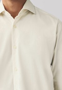 Camisa de botones de crema clara con una tela texturizada, cuello tipo spread y cuatro botones en la parte delantera. Apariencia suave, diseño ajustado.