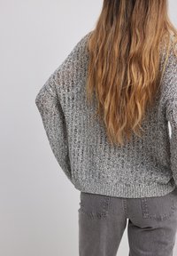 Cheveux longs et ondulés vus de dos, une personne portant un pull gris en maille ajourée et un jean gris en denim sur un fond uni.