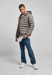 Grå pufferjakke med hætte, quiltet design og frontlynlås; lagdelt over en hvid skjorte, parret med blå jeans og hvide sneakers.