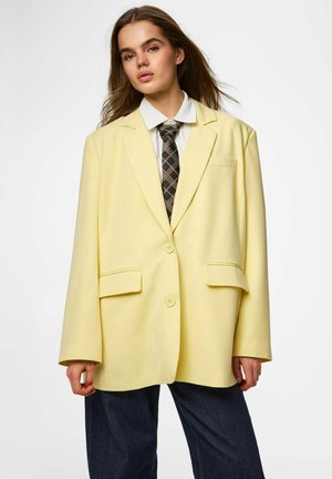 Jonge vrouw in een oversized bleekgele blazer, wit overhemd met kraag, een patroon stropdas en donkere, wijde broek tegen een witte achtergrond.