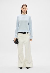 Světle modrý svetr, krémové kalhoty s širokými nohavicemi, černý opasek a černá kabelka. Modelka stojí v dlouhých rukávech s minimálními detaily.
