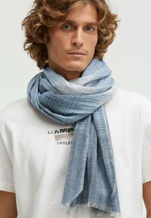 Young man with curly blonde hair wearing a white graphic t-shirt and a soft blue patterned scarf wrapped around his neck.