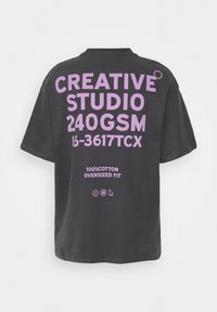 Crna oversized majica od 100% pamuka, s velikim ljubičastim natpisom na leđima i malim grafičkim akcentima blizu izreza.