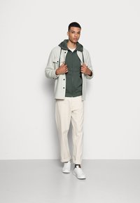 Chemise boutonnée gris clair sur un hoodie vert, pantalon crème et baskets blanches, créant un look décontracté en superposition.