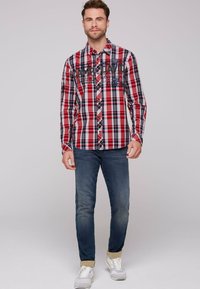 Rot und blau kariertes Langarmhemd mit Knopfleiste, mit Textdetails auf der Brust, kombiniert mit blauen Jeans und weißen Turnschuhen.