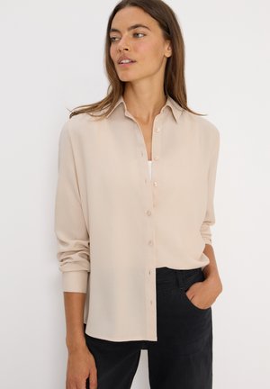 Beige Hemd mit Kragen und langen Ärmeln, aus leichtem Stoff gefertigt. Kombiniert mit dunklen Hosen.