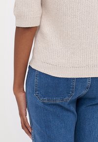 Haut beige à manches courtes en maille avec une texture côtelée, associé à un jean bleu en denim avec des poches arrière et une coupe droite.