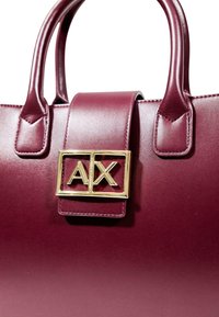 Borsa in pelle bordeaux con due manici superiori, caratterizzata da una chiusura rettangolare dorata con "AX" al centro. Texture liscia e design strutturato.