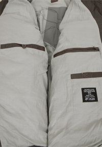 Helles graues Jackenfutter mit braunen Akzenten, ausgestattet mit zwei Reißverschlusstaschen und einem dekorativen Knopf. Enthält ein Pflegeetikett der Marke.