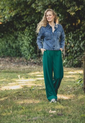 Spijkerjasje met twee voorzakken, gedragen met losse groene broeken en lichtgekleurde sneakers, in een natuurlijke buitensetting.
