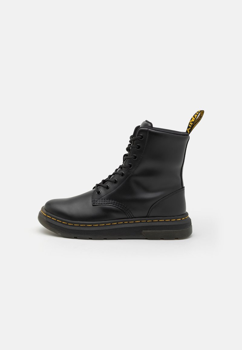 Botte en cuir noire à lacets avec semelle épaisse et coutures jaunes, avec une languette jaune à l’arrière avec texte noir.