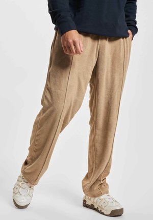 Person wearing beige fleece pants paired with white sneakers and a dark long-sleeve top, standing with one hand in pocket.