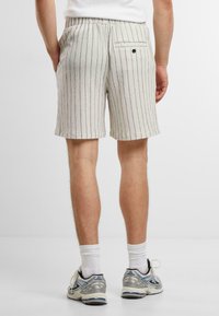 Bermudas gris claro, texturizadas, con rayas verticales, cinturilla elástica y un bolsillo trasero con detalle de botón. Zapatillas deportivas visibles.