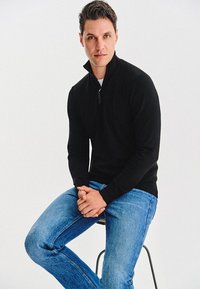 Czarny sweter zapinany na zamek z wysokim kołnierzem, wykonany z dzianiny, zestawiony z jasnoniebieskimi dżinsami. Model siedzi na stołku ze złożonymi rękami.