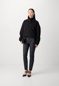 Černý oversized polo s vysokým límcem, tmavé kalhoty a černé špičaté podpatky s zlatými kulatými akcenty.