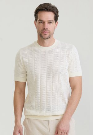 Man with short brown hair wearing a short-sleeve white knit sweater and light beige pants against a plain light background.