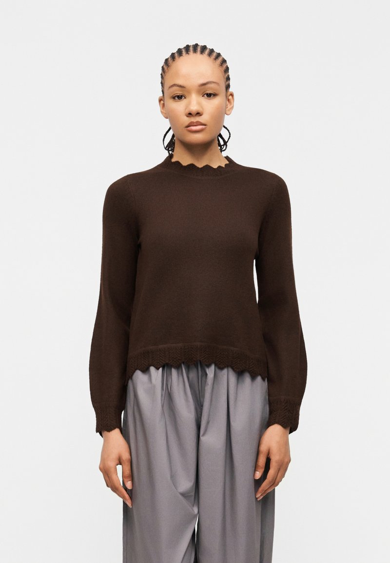 Γυναίκα με πλεξούδες φοράει σκούρο καφέ πουλόβερ με μακριά μανίκια και κυματιστά τελειώματα, καθώς και χαλαρά γκρι παντελόνια, στέκεται μπροστά σε απλό φόντο.