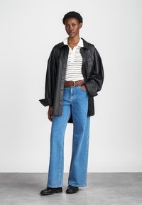 Black leather oversized shirt, white striped polo, high-waisted blue denim jeans, brown woven belt, black ankle boots, standing pose.