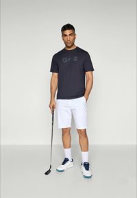 Man in black t-shirt, white shorts, holding golf club, standing on plain background, wearing white socks and white golf shoes with blue laces.