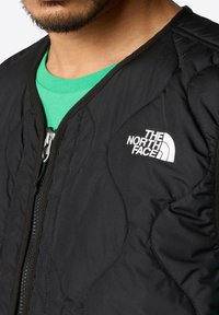 Black quilted jacket featuring a high collar and front zipper. The left chest displays a white logo. A green shirt is visible underneath.