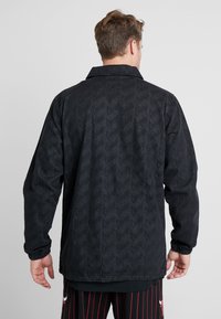 Man med svart jacka med subtilt upprepande textmönster och svarta basketshorts med röda ränder och en logo på baksidan.