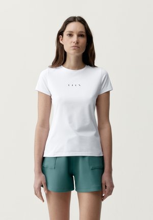 Žena s dlhými hnedými vlasmi, oblečená v bielom tričku s malým čiernym potlačou fáz mesiaca a v tyrkysových šortkách, stojaca pred jednofarebným pozadím.