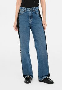 Jeans in denim blu con gambe larghe, un accento in tessuto nero sfrangiato sulle cuciture laterali e un classico design a cinque tasche.