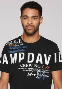 Camiseta de algodón negra con gran texto blanco "CAMP DAVID" y acentos azules. Cuello redondo con detalles impresos en el hombro.