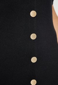 Gros plan sur un tissu noir avec une rangée verticale de cinq boutons ronds en or, également espacés, le long du côté.