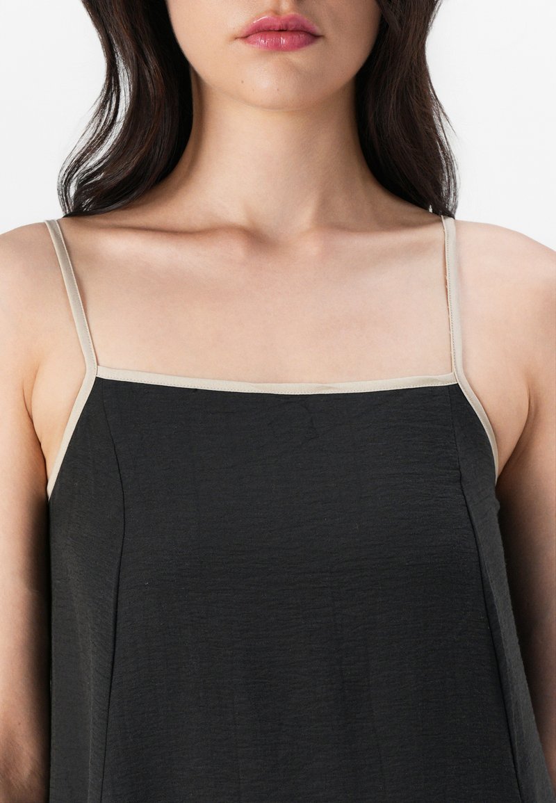 Zwarte mouwloze top met dunne beige schouderbandjes, vervaardigd uit een zachte, gestructureerde stof en met een rechte halslijn. Geen patronen of versieringen.