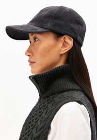 Donker corduroy pet met een gebogen rand, voorzien van een gestructureerd patroon. Gecombineerd met een donkere gebreide mouwloze trui en een wit shirt eronder.