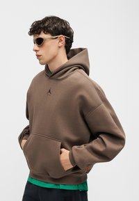 Jeune homme portant un sweat à capuche marron avec le logo Air Jordan et des lunettes de soleil, mains dans la poche avant, regardant à gauche sur un fond uni.