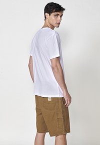 T-shirt bianco a maniche corte e pantaloni cargo marroni. I pantaloni hanno diverse tasche e una toppa sul retro, mostrati da un leggero angolo.