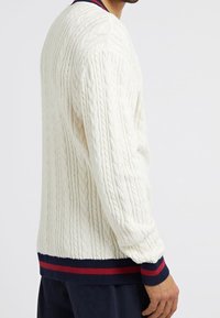 Homme portant un pull en maille torsadée blanche avec des poignets et un ourlet rayés bleu marine et rouges, associé à un pantalon foncé, vu de profil.