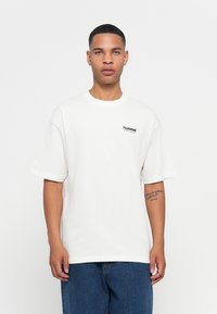 Witte katoenen t-shirt met korte mouwen en ronde halslijn. Bevat een klein zwart "hummel" logo op de borst. Draagt samen met blauwe denim jeans.