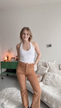 Mlada žena u bijelom topu na vezanje i smešnim hlačama od kordura stoji u udobnoj dnevnoj sobi s zelenim ormarom i bijelom sofom.