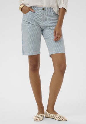 Nő térdig érő kék-fehér csíkos rövidnadrágban, fehér blúzban, arany karkötővel, gyűrűkkel és bézs színű fonott balerinacipőben.