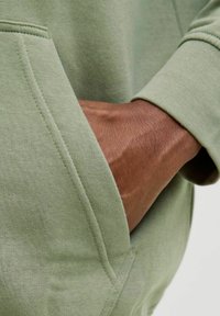 Grön hoodie i bomull med framficka; har en mjuk textur och detaljer vid muddarna; hand som vilar i fickan.