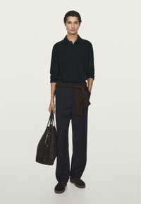 Svart långärmad polo, mörkbrun tröja knuten i midjan, lösa marinblå byxor och en mörk väska. Modellen håller en mörk väska med två handtag.